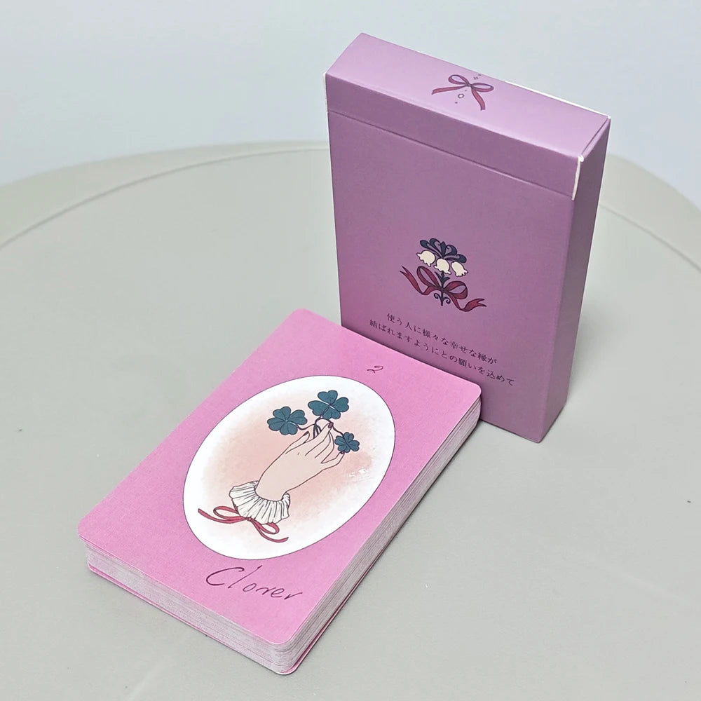 Ribbon Lenormand — pink deck with Clover card (hand holding four-leaf clovers) beside pink rigid box with lily of the valley and ribbon motif and Japanese text
