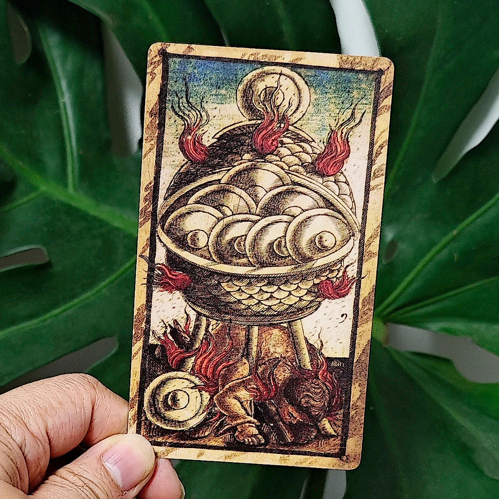 Sola Busca Tarot — 10 of Wands card held up: a large serpent-scaled cauldron overflowing with flaming wands atop a lion-paw pedestal, with a small lion figure below; held against monstera leaves