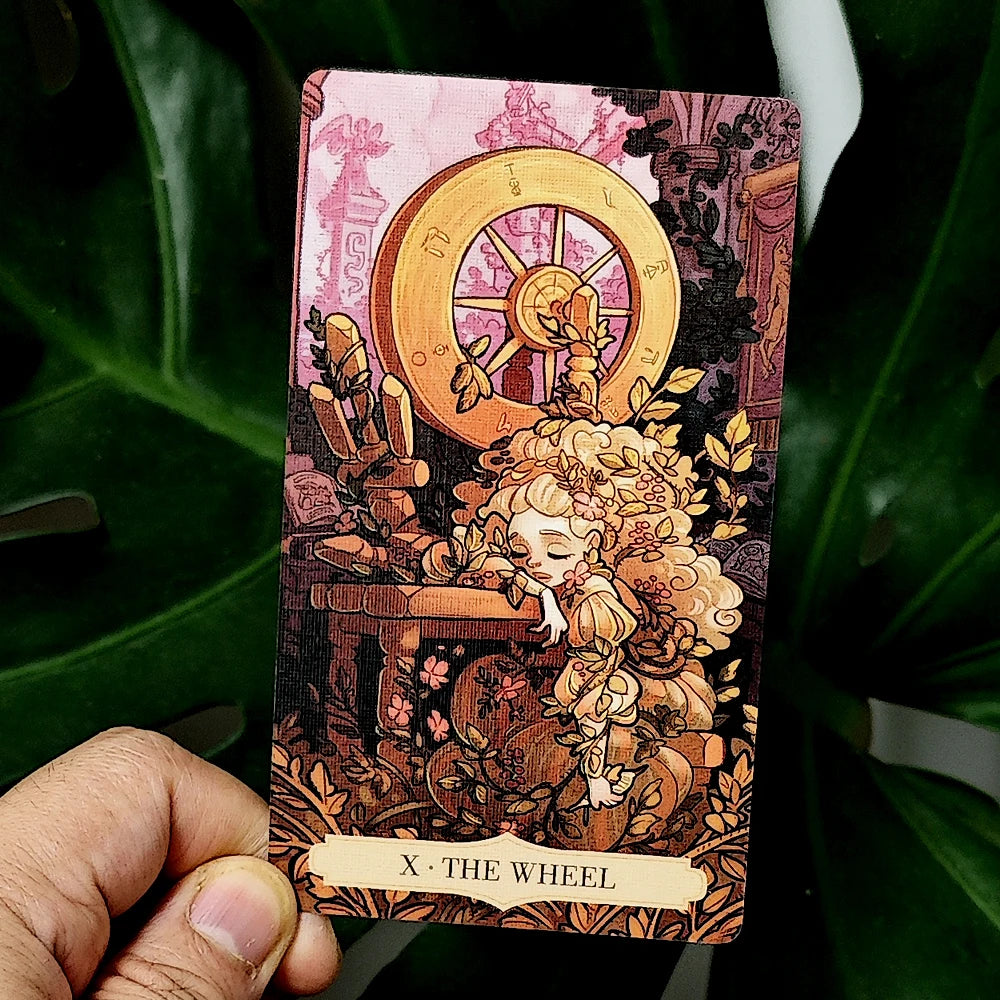 Once Upon a Time Tarot — X The Wheel card: dreaming blonde girl amid golden spinning wheel, flowering vines, and autumn leaves in a pink baroque ruin; warm amber and rose palette