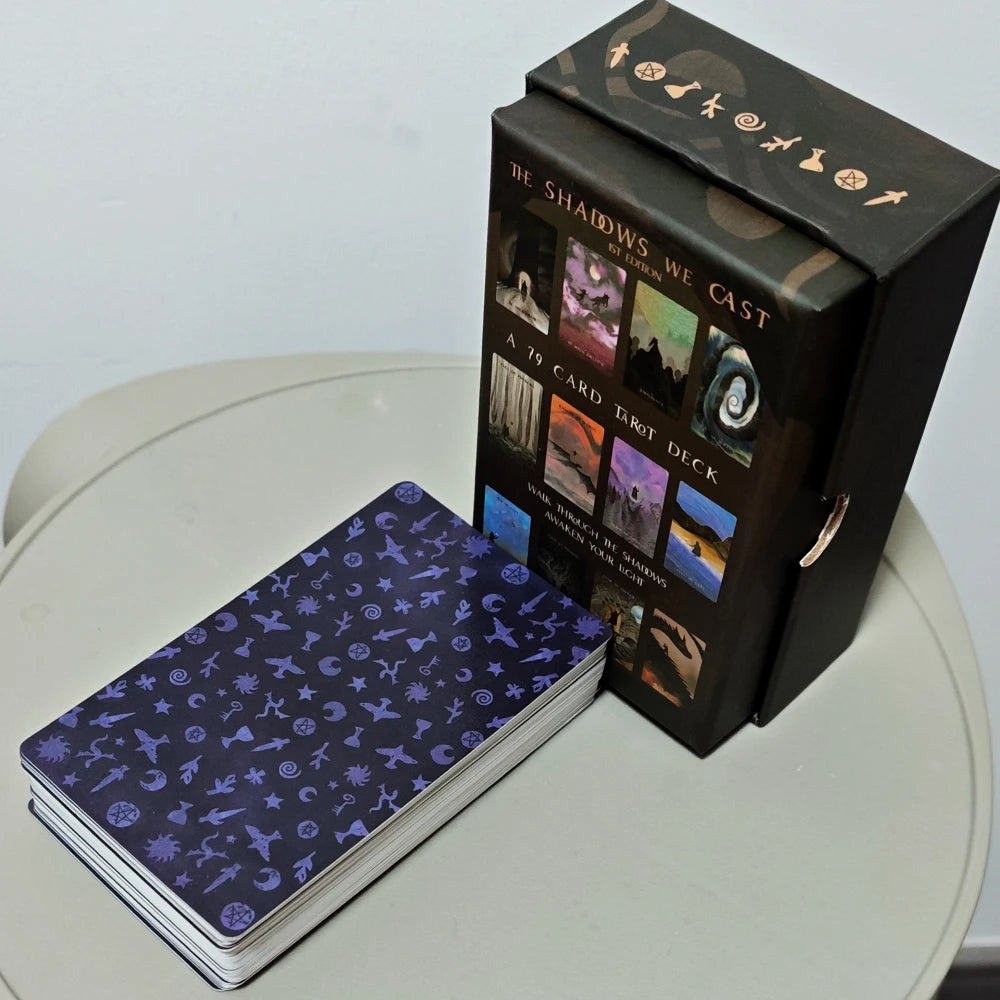 The Shadows We Cast Tarot — 79-card deck beside rigid box showing copper esoteric symbols; midnight blue card backs with patterned esoteric icons