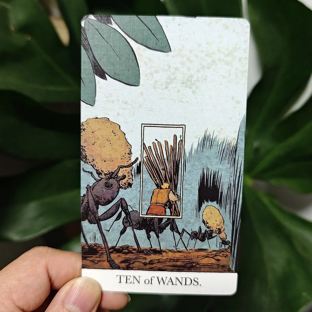 The Unveiled Tarot — Ten of Wands card held up showing ants carrying a card of a figure burdened with wands, against a monstera plant background