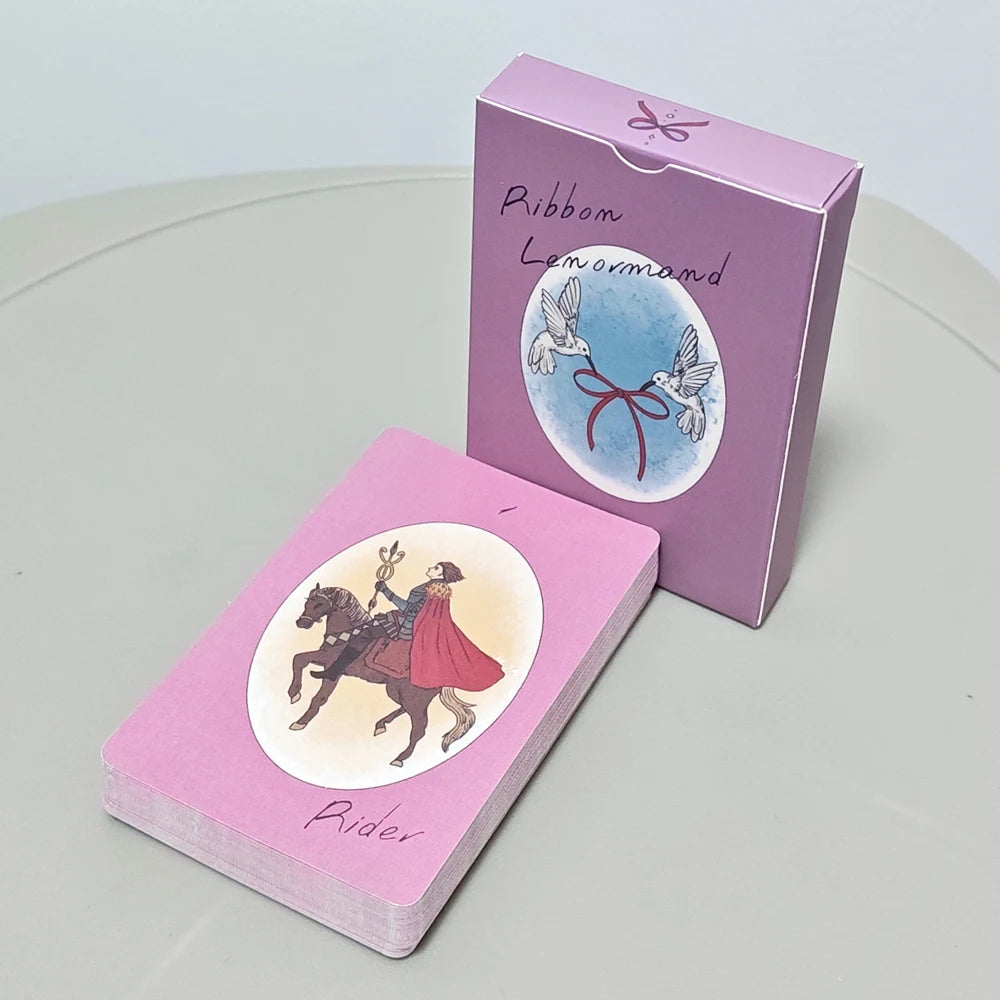 Ribbon Lenormand — Rider card on top of full deck beside matte pink-purple rigid box showing Birds with crimson ribbon on front panel