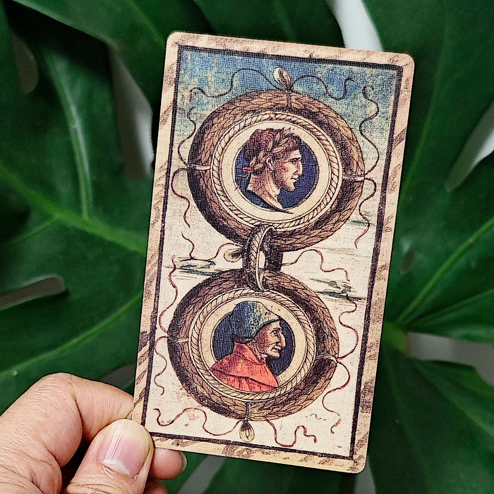 Sola Busca Tarot — 2 of Coins card held up: two Roman portrait medallions in profile (one in a laurel wreath, one in a red cap) framed by intertwined ouroboros-rope borders, held against monstera leaves